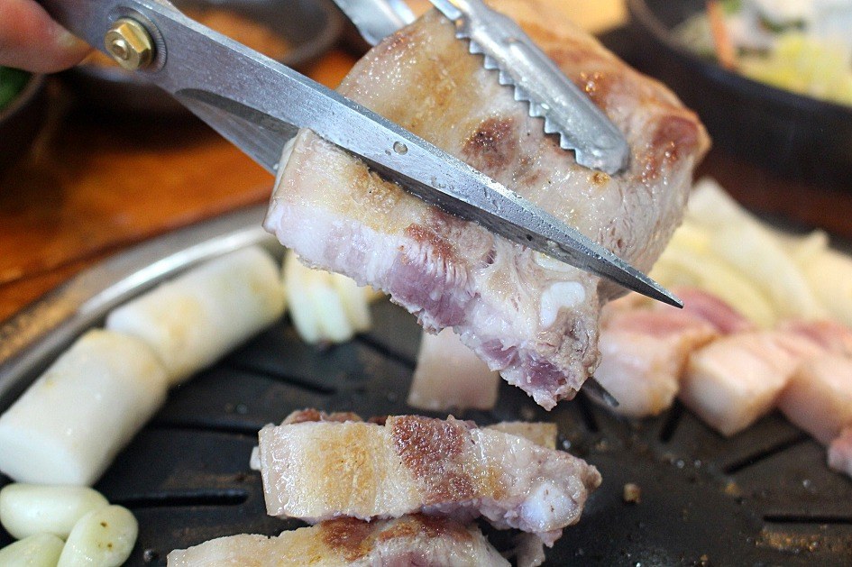 Kitchen scissors and tongs cutting grilled pork belly (samgyeopsal) into pieces on a Korean BBQ grill