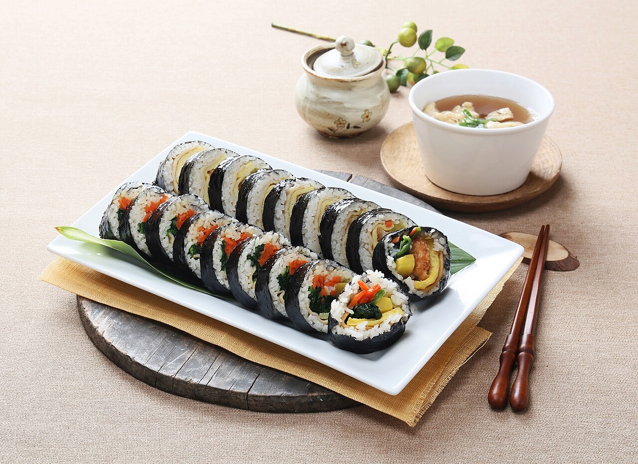 Two rolls of sliced Korean gimbap served on a white plate with a bowl of soup
