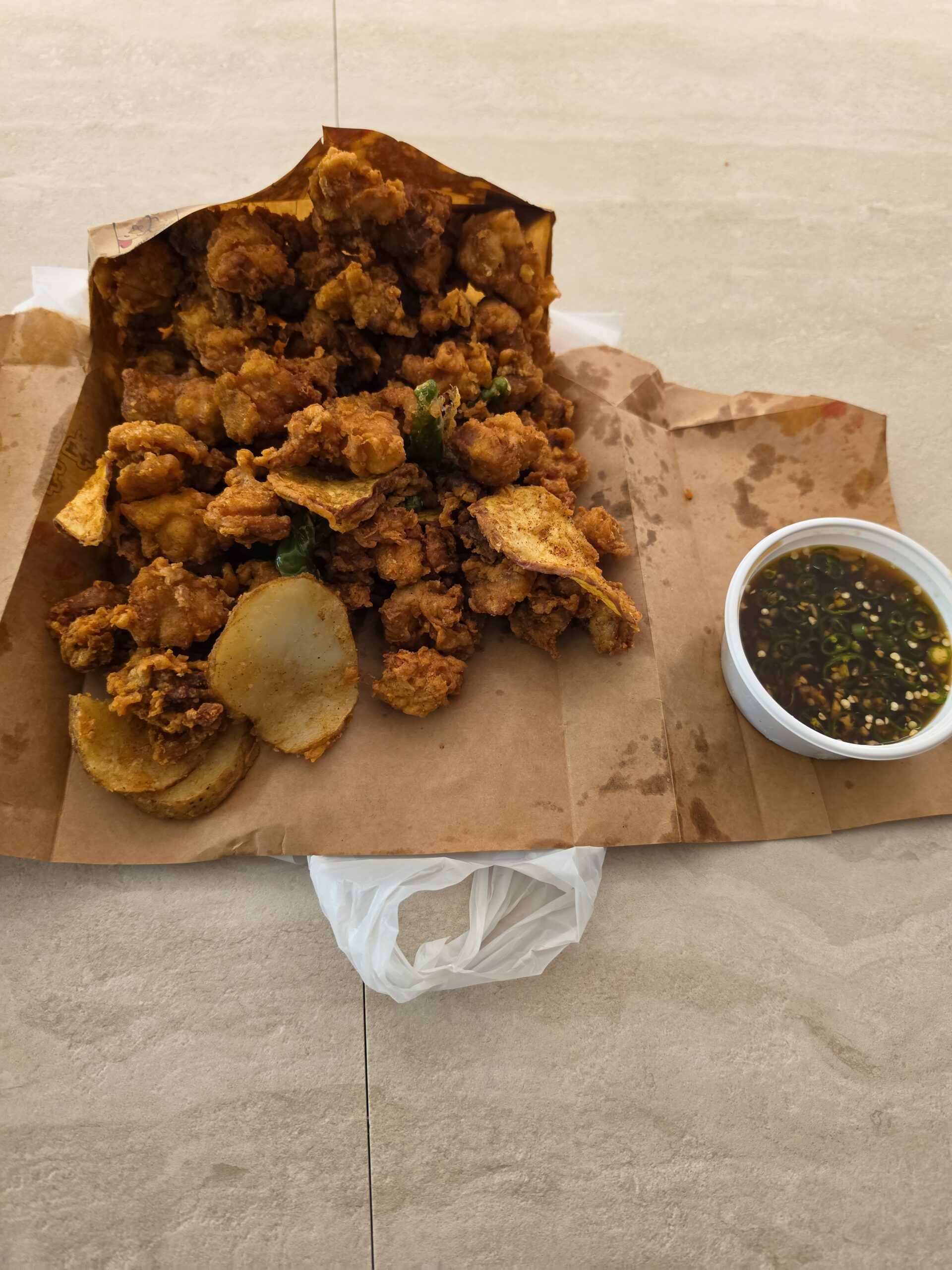 Korean boneless fried chicken served on its opened brown paper takeaway wrapping, with potato slices and dipping sauce