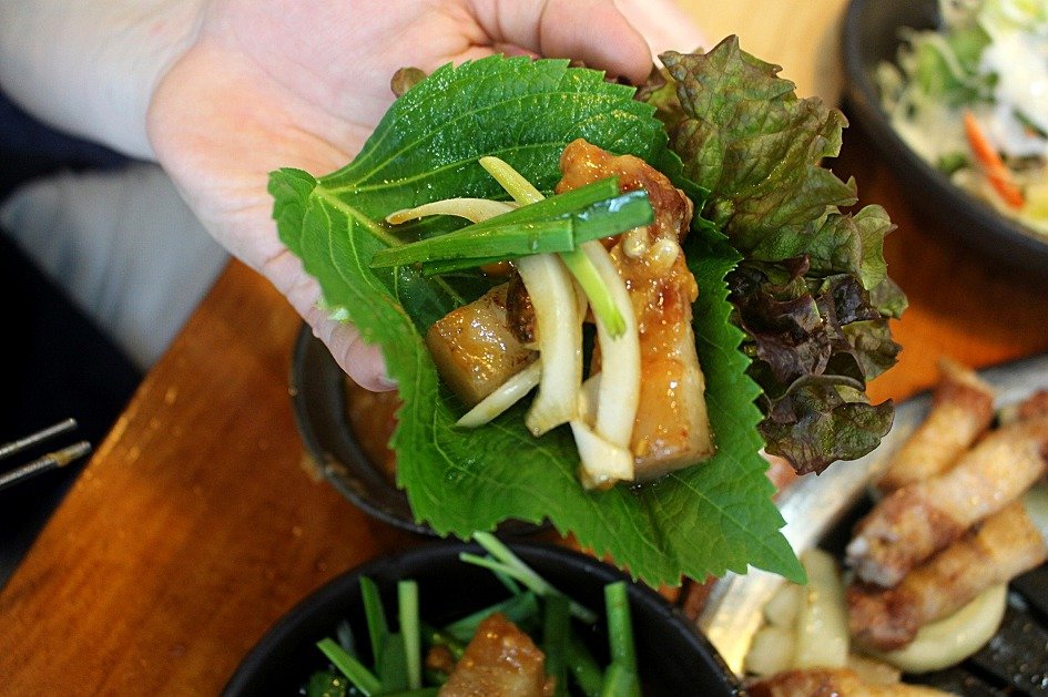 Hand holding a ssam wrap of grilled pork belly in a perilla leaf at a Korean BBQ restaurant