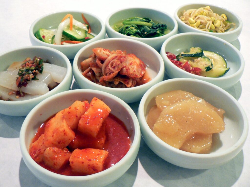 Eight small white bowls of Korean banchan side dishes, including cucumber, spinach, bean sprouts, and radish kimchi.