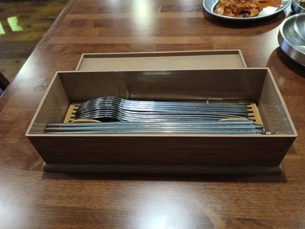 An open tan utensil box on a wooden Korean restaurant table, holding rows of flat metal chopsticks and long-handled spoons.